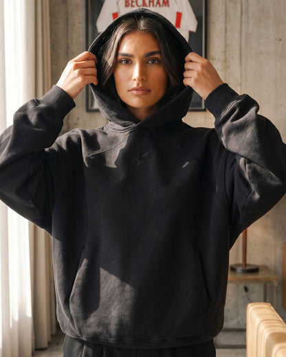 A woman wearing a black oversized pullover hoodie with a small embroidered star logo on the chest, holding up the hood. The setting is a rustic indoor room with concrete walls and natural light.