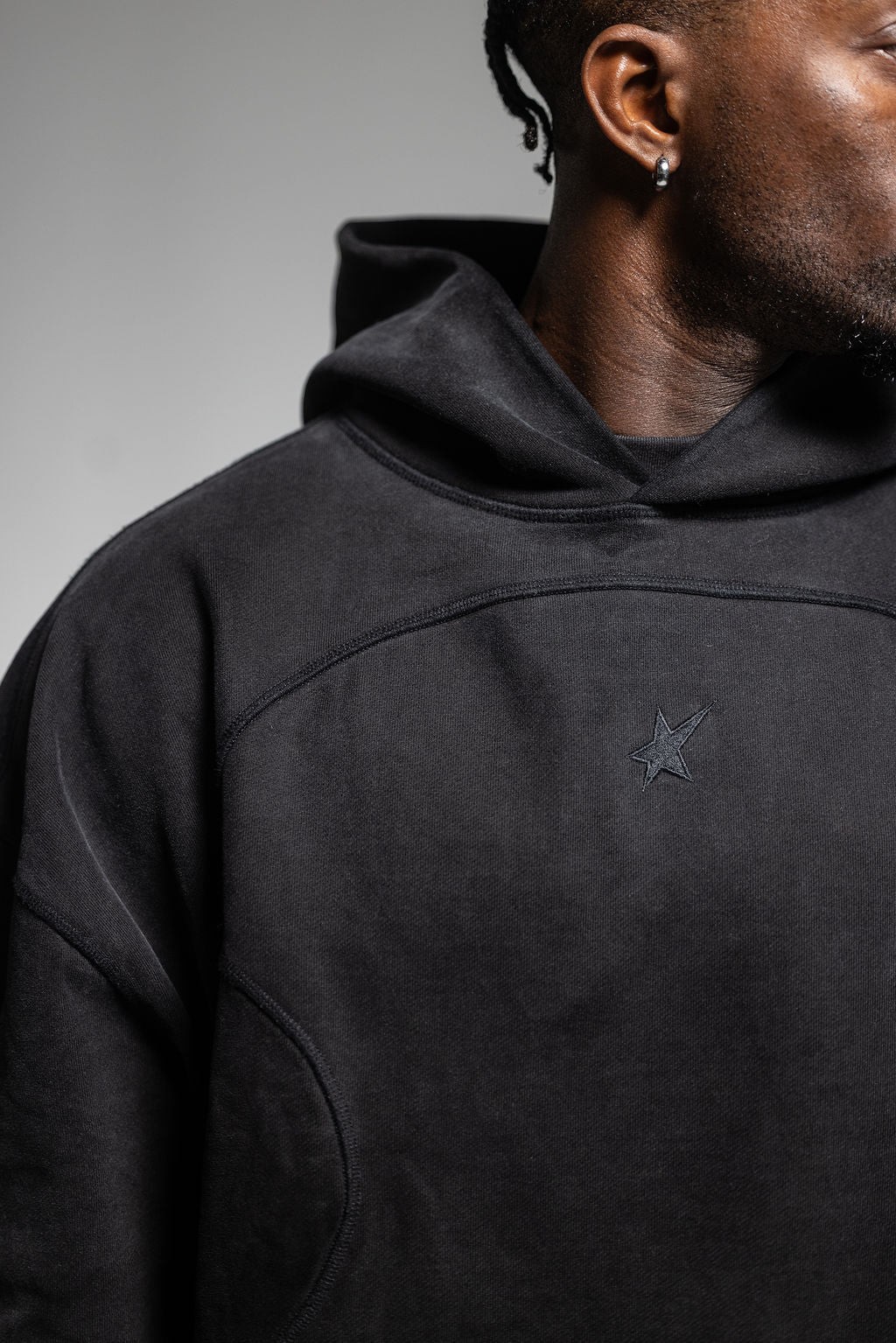 A close-up of a person wearing a black hooded sweatshirt against a neutral gray background. The hoodie features a black embroidered star logo.
