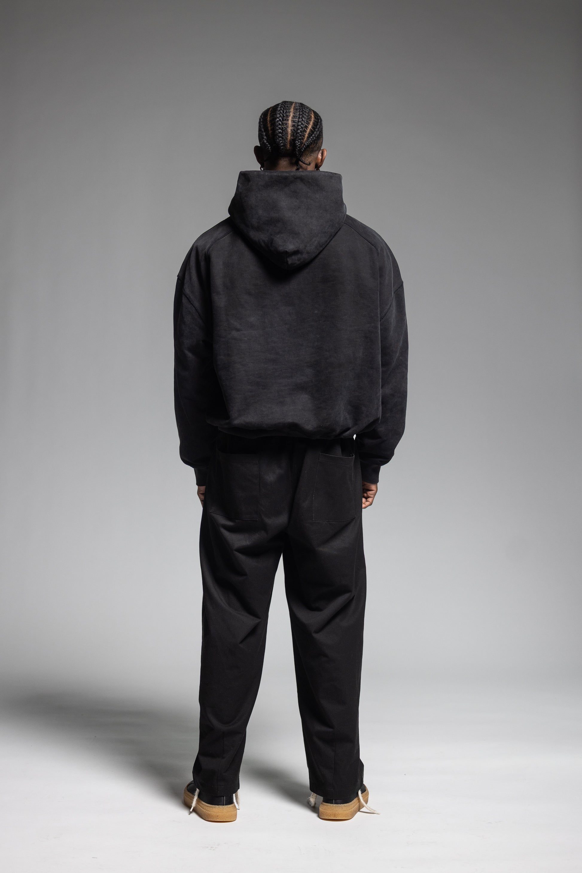 A model viewed from behind wearing Trew Collections Black on Black Hoodie with black trousers, standing against a neutral gray studio background.