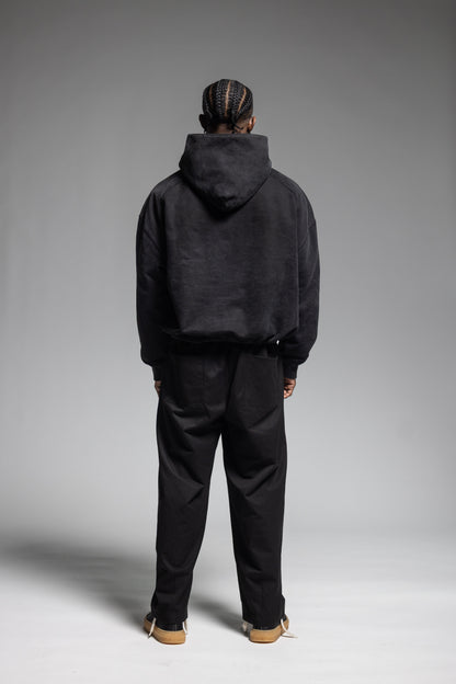 A model viewed from behind wearing Trew Collections Black on Black Hoodie with black trousers, standing against a neutral gray studio background.