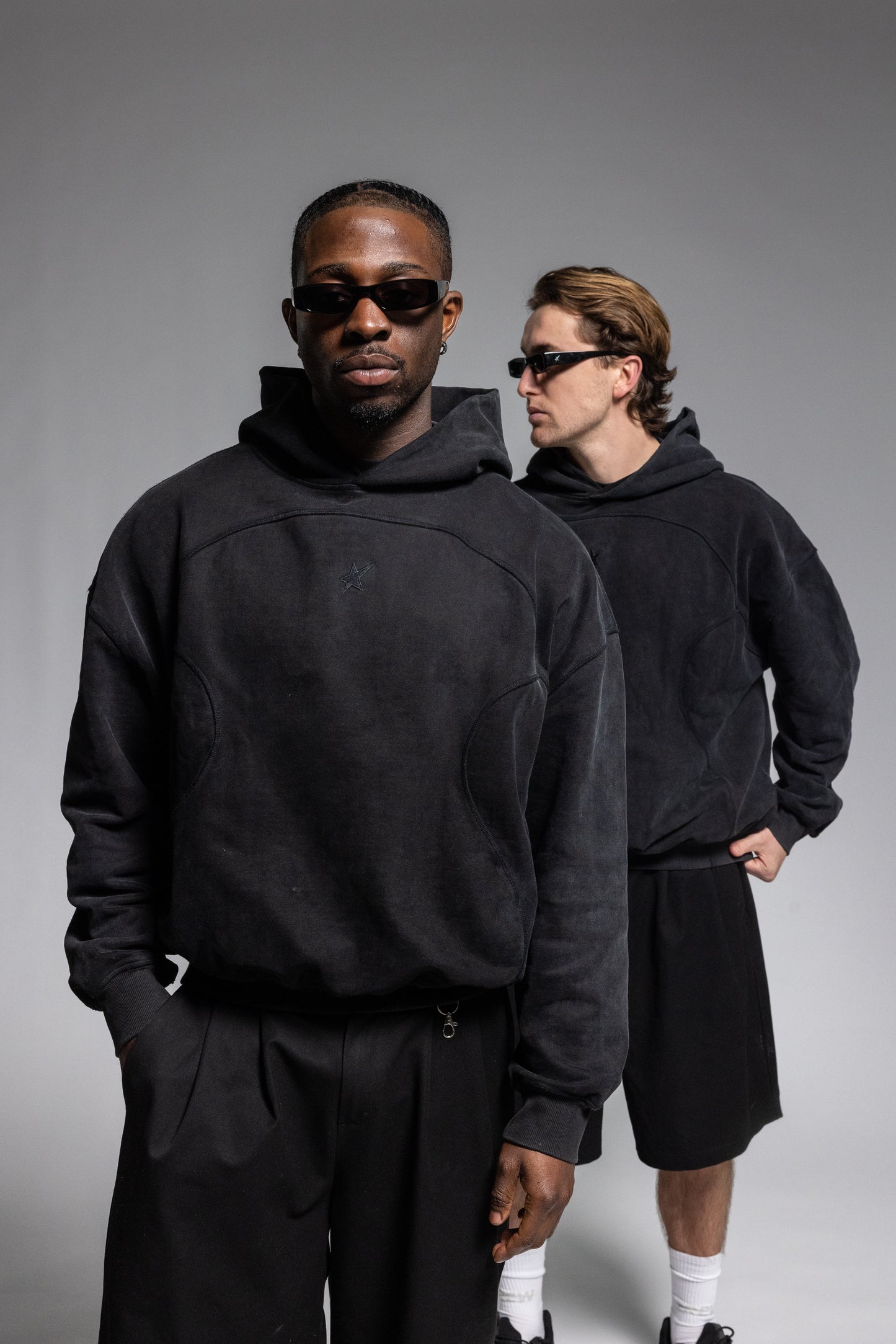 Two models wearing Trew Collections Black on Black Hoodie, styled with black sunglasses, white socks, and black sneakers against a solid gray studio background.