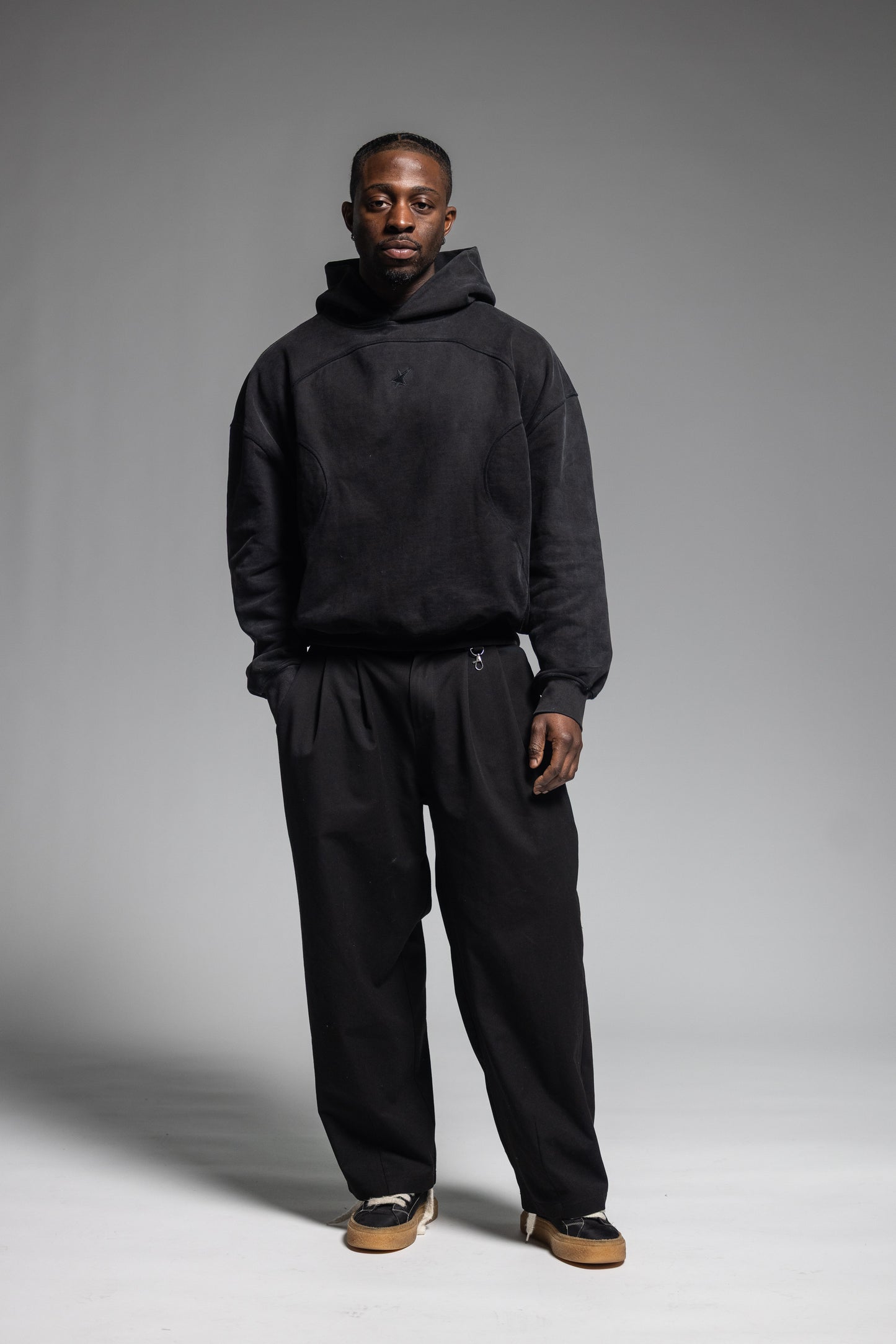 A man modeling Trew Collections Black on Black Hoodie with black trousers against a neutral gray studio background.