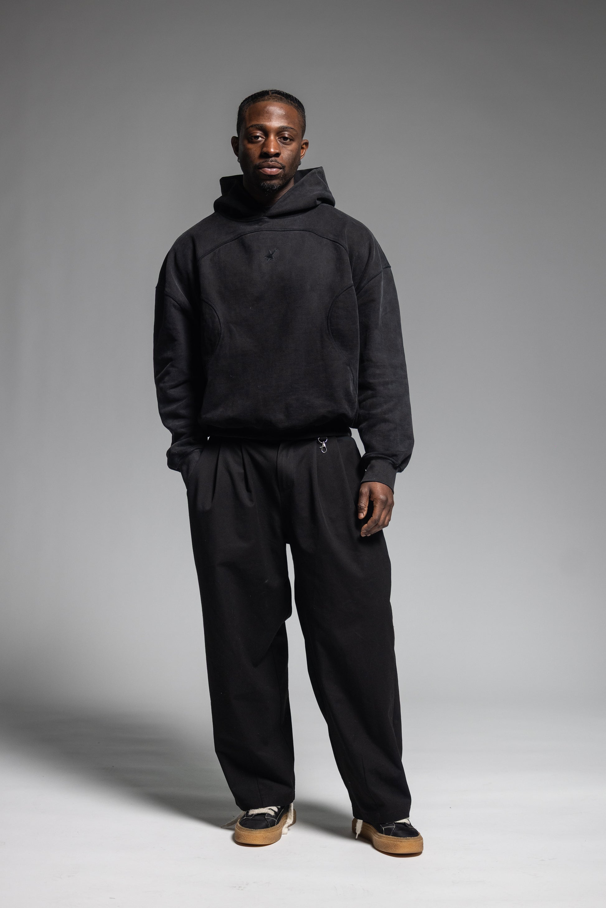 A man modeling Trew Collections Black on Black Hoodie with black trousers against a neutral gray studio background.
