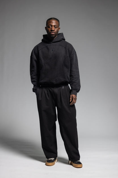 A man modeling Trew Collections Black on Black Hoodie with black trousers against a neutral gray studio background.