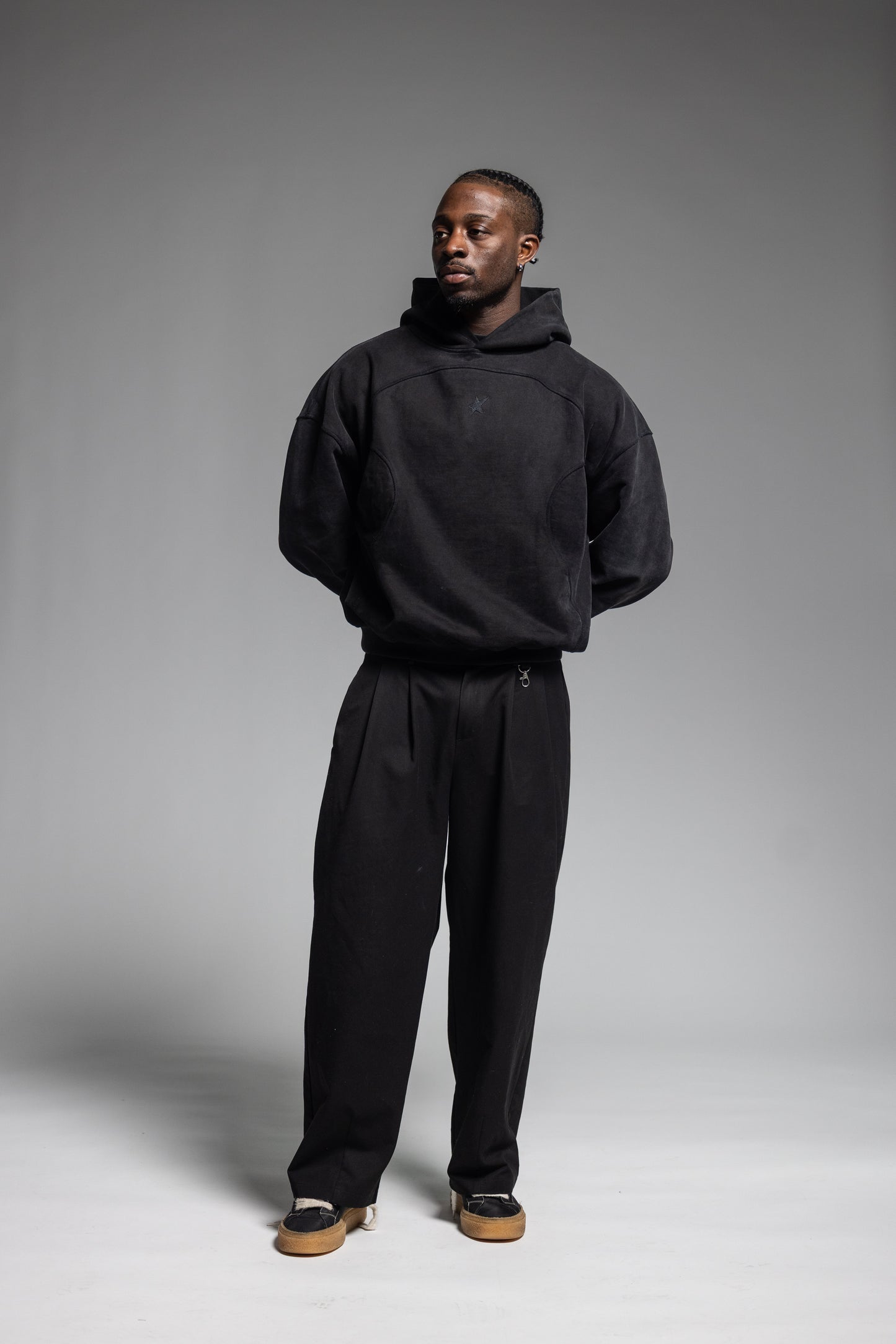 A man modeling Trew Collections Black on Black Hoodie and black trousers against a neutral gray studio background.