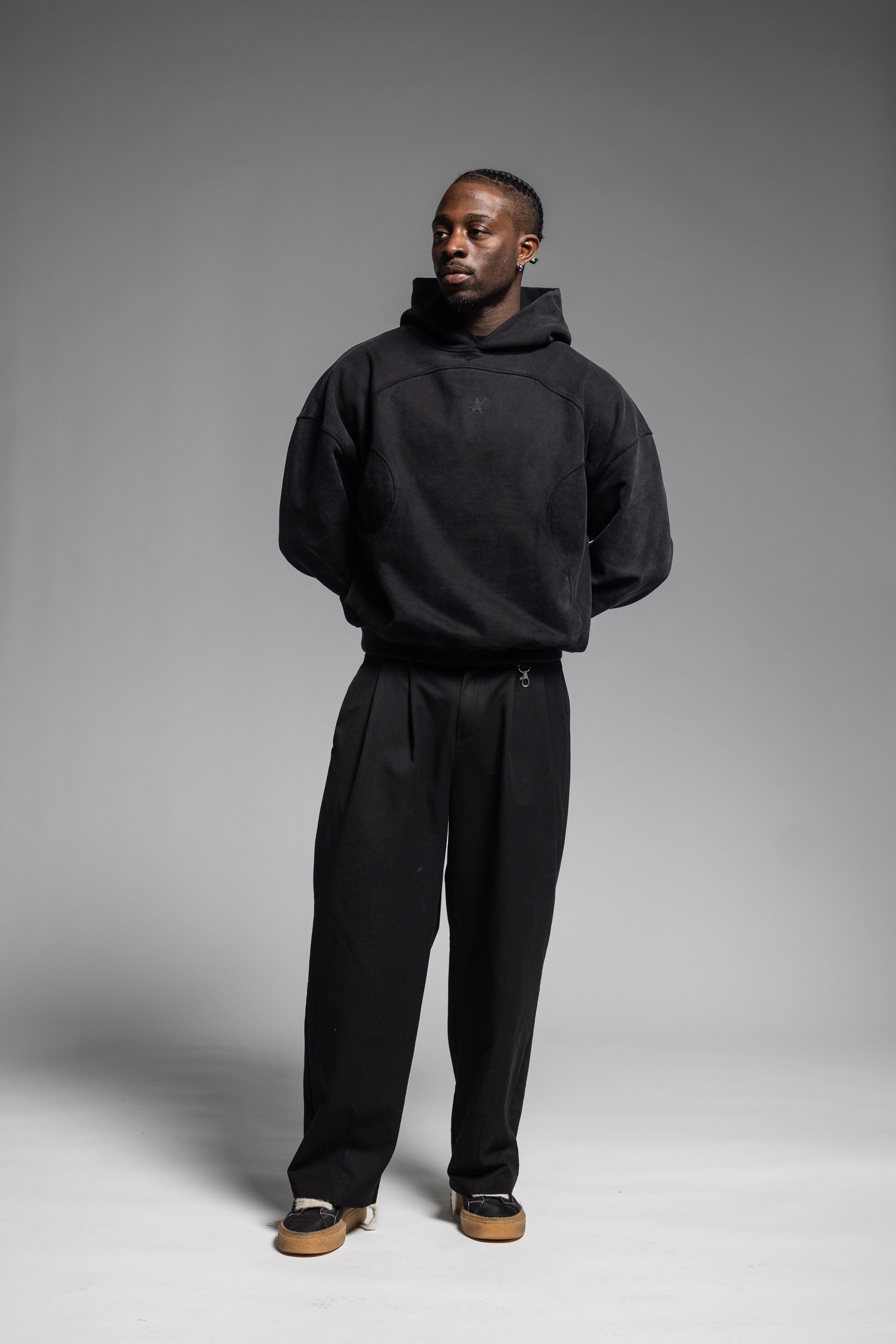 A man modeling Trew Collections Black on Black Hoodie and black trousers against a neutral gray studio background.