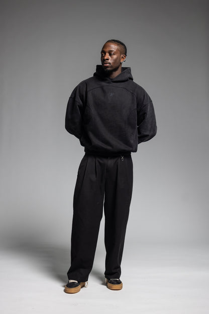 A man modeling Trew Collections Black on Black Hoodie and black trousers against a neutral gray studio background.