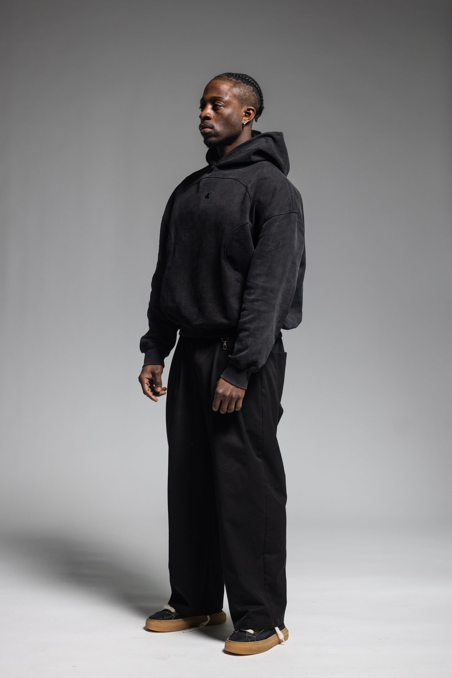 A man wearing Trew Collections Black on Black Hoodie with black trousers, standing in profile against a neutral gray studio background.