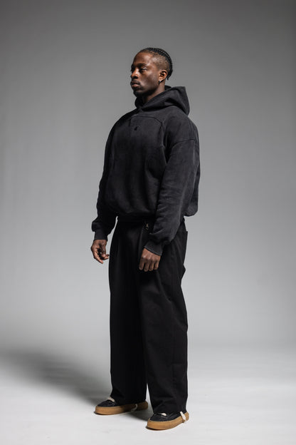 A man wearing Trew Collections Black on Black Hoodie with black trousers, standing in profile against a neutral gray studio background.