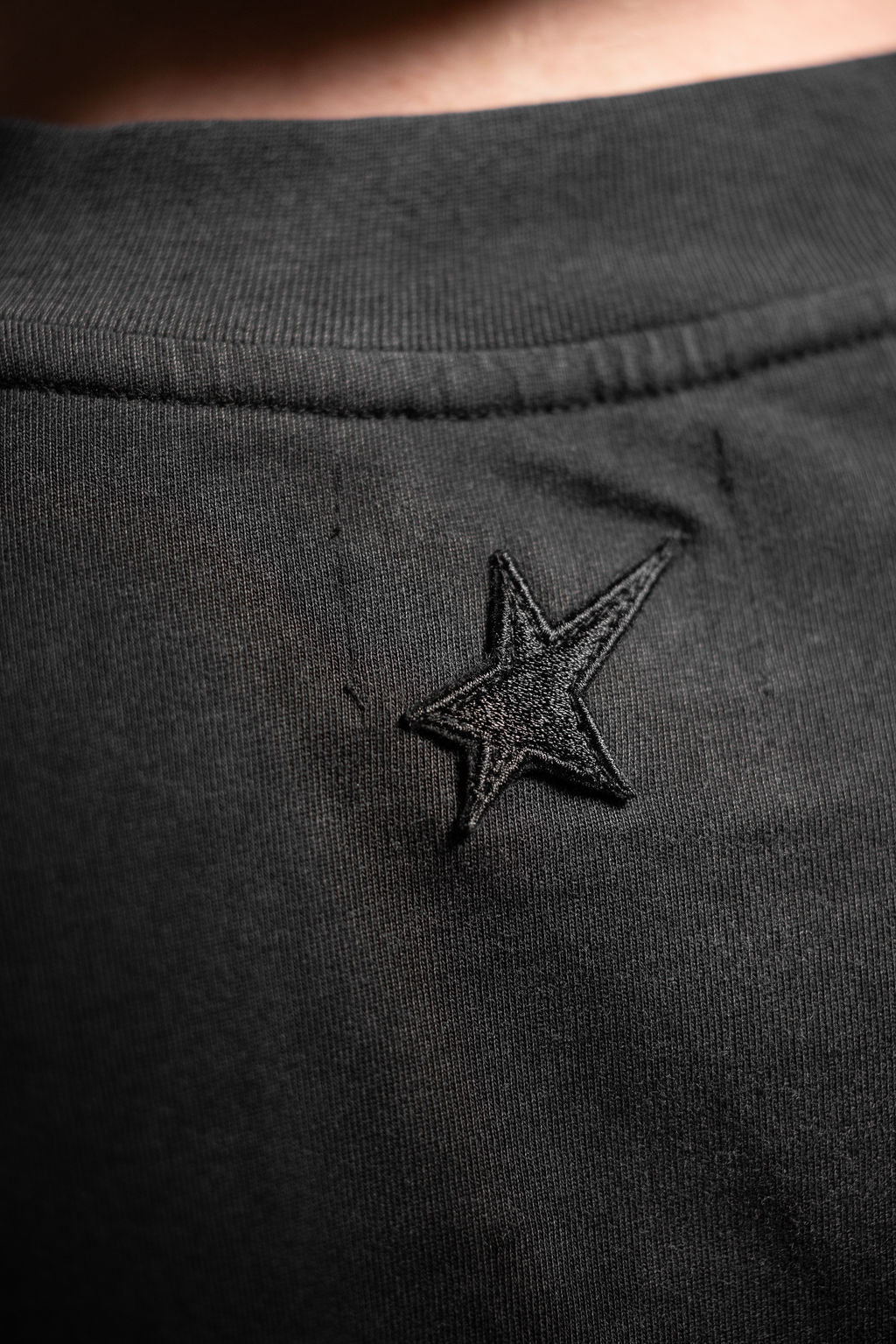 Close-up of a black cotton Tee with an embroidered black star logo on the chest.
