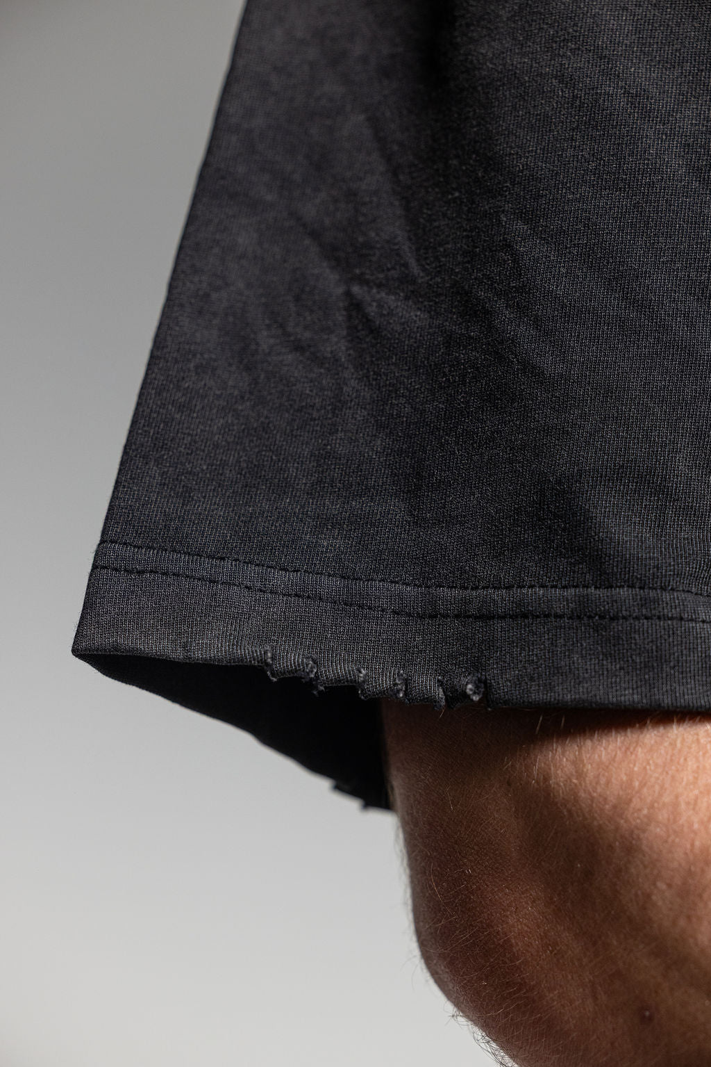 Close-up of a black short-sleeve shirt sleeve with raw, frayed hems, worn on a person's arm against a neutral gray background.