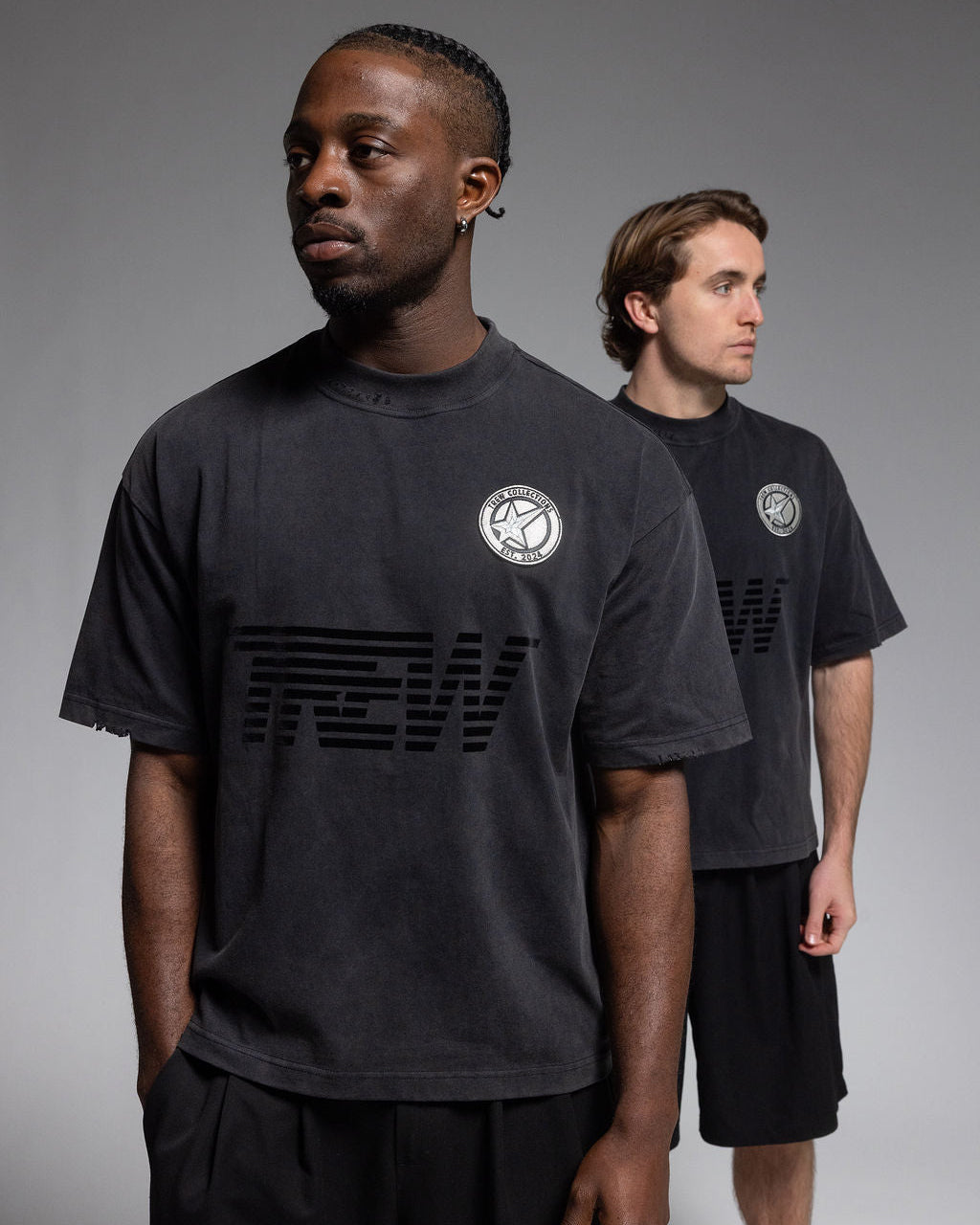 Two male models wearing dark charcoal gray graphic t-shirts with a circular crest logo on the chest and a large tonal graphic across the front, posed against a neutral gray studio background.