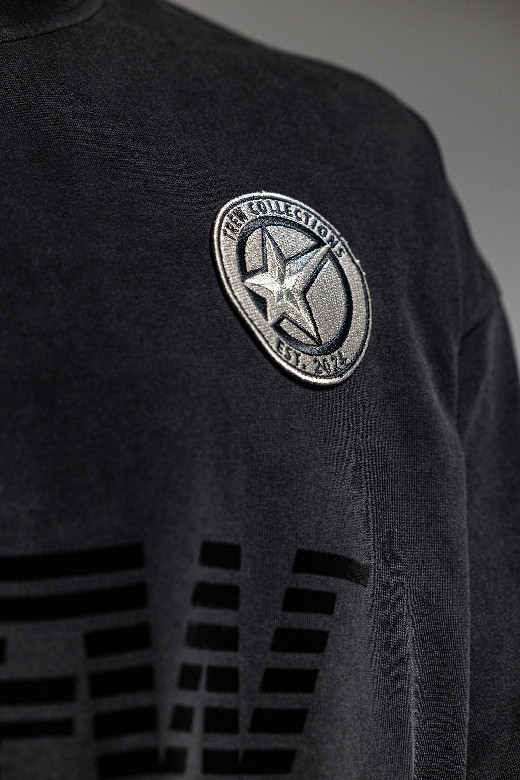 Close-up of washed-out black Tee featuring embroidered logo patch on chest.