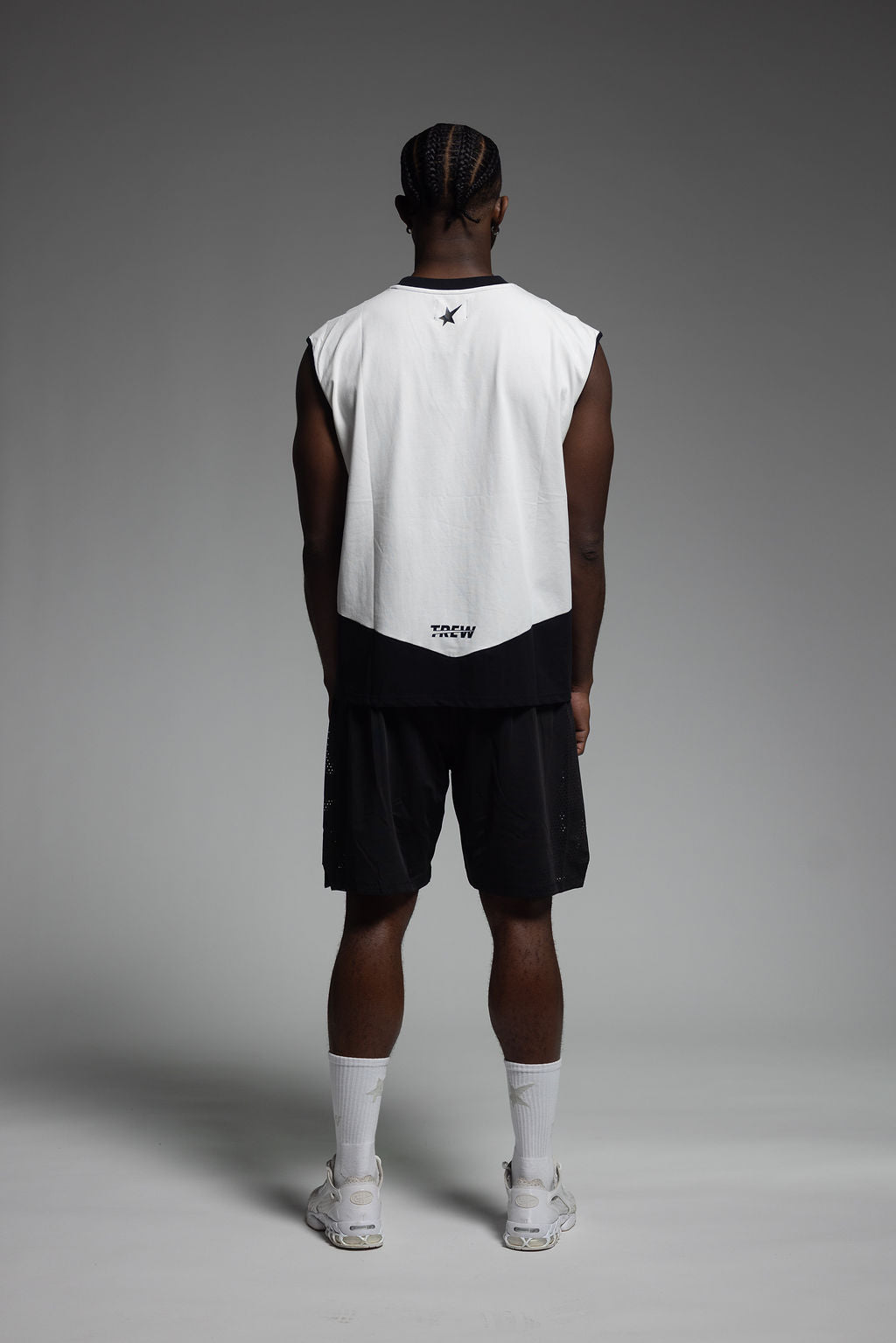 A man stands with his back to the camera wearing a sleeveless white and black athletic jersey, black shorts, white socks, and white sneakers against a gray studio background.