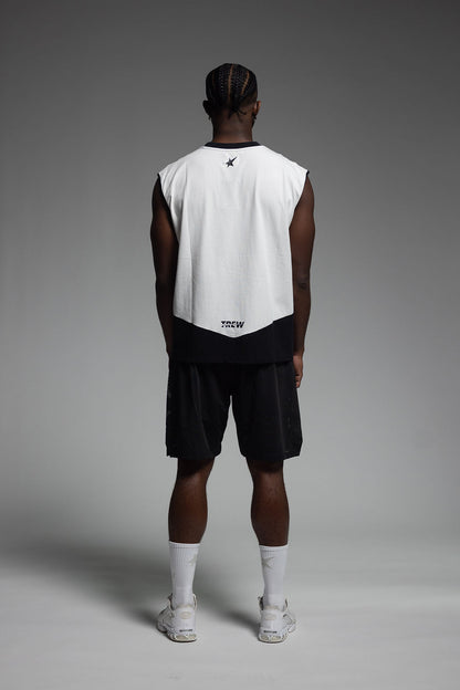A man stands with his back to the camera wearing a sleeveless white and black athletic jersey, black shorts, white socks, and white sneakers against a gray studio background.