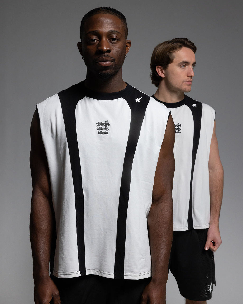 Two models wearing sleeveless white athletic jerseys with black side panels and black shorts, standing against a dark gray studio background.