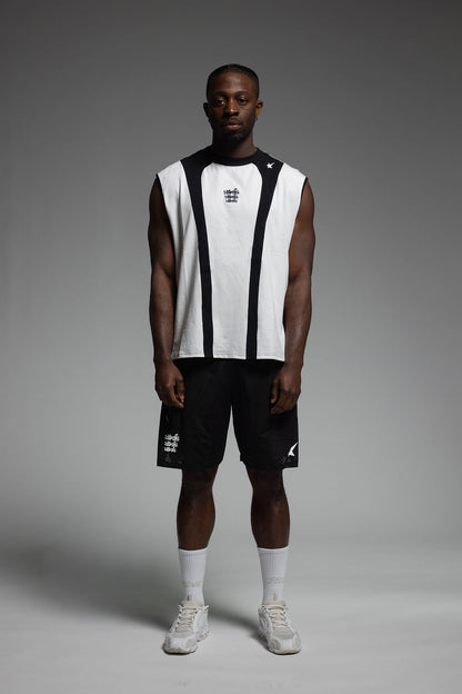 A model stands against a dark gray studio background wearing a sleeveless white and black athletic top, black athletic shorts, white socks, and white sneakers.