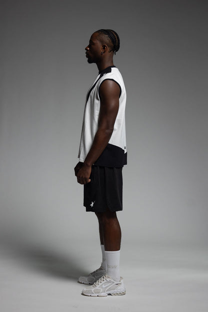 A man in profile wearing a white training singlet with black trim, black shorts, white socks, and white sneakers, standing against a smooth gray studio background.