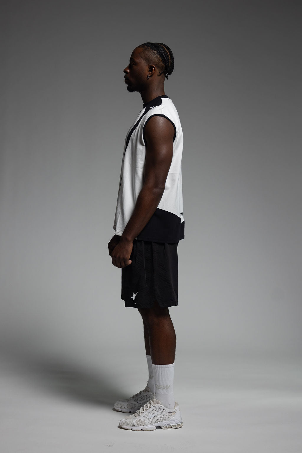 A man in profile wearing a white sleeveless athletic top with black trim, black shorts, white socks, and white sneakers, standing against a smooth gray studio background.
