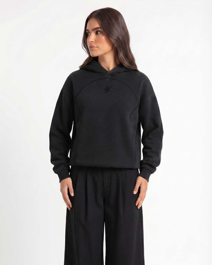 A woman modeling Trew Collections back on Black Hoodie, paired with black wide-leg sweatpants, against a plain light gray studio background.
