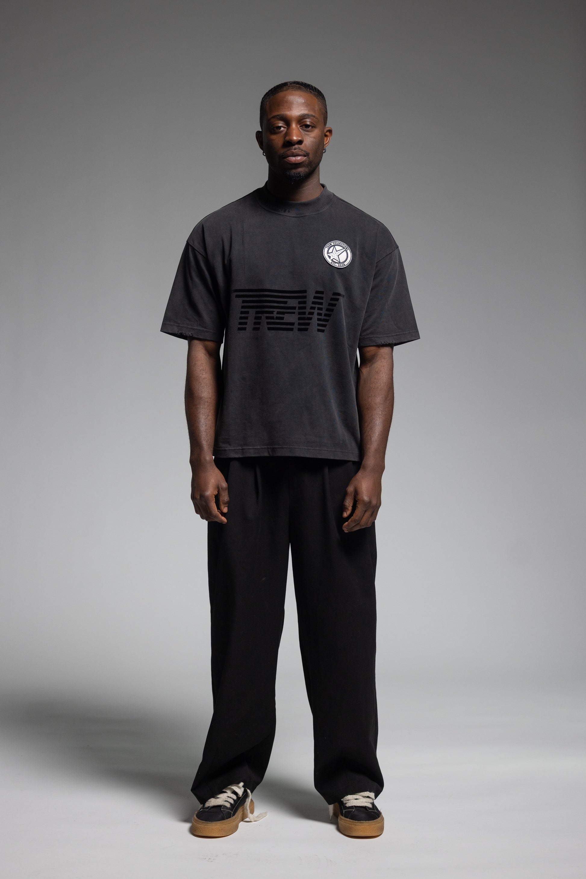 A man stands against a gray studio background wearing Trew Collections Black on Black Tee, black pants, and sneakers.