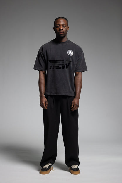 A man stands against a gray studio background wearing Trew Collections Black on Black Tee, black pants, and sneakers.