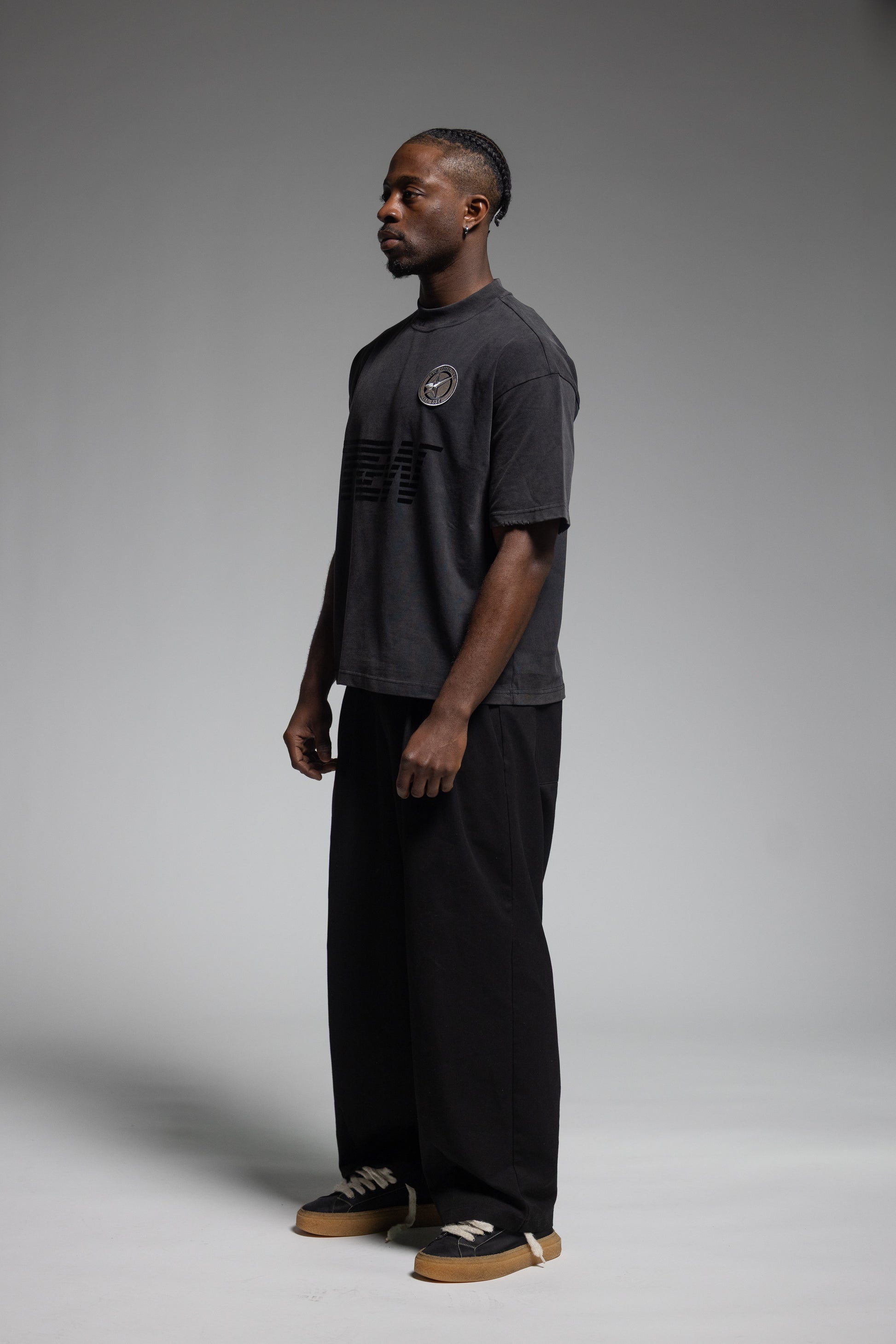 A man stands in profile wearing Trew Collections Back on Black Tee, paired black wide-leg pants and sneakers, against a neutral gray studio background.