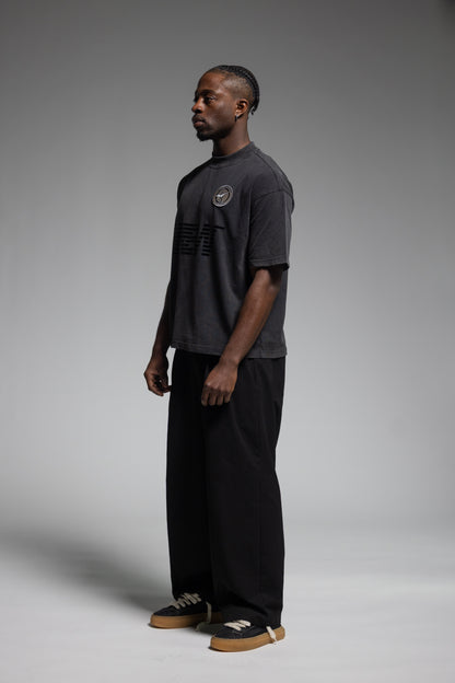 A man stands in profile wearing Trew Collections Back on Black Tee, paired black wide-leg pants and sneakers, against a neutral gray studio background.